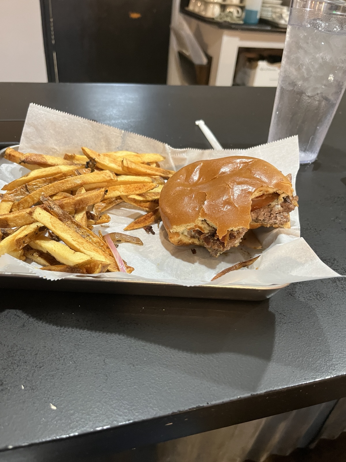 A partially eaten burger with a glossy bun sits in a paper-lined tray alongside seasoned fries, placed on a dark surface with a glass of water and background elements visible.