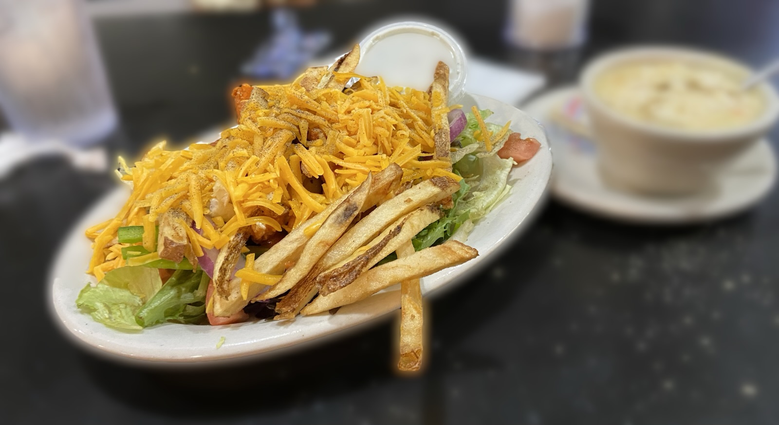 A plate of salad topped with shredded cheese, crispy fried items, and a side of dressing sits on a dark table, with a bowl of soup in the background.