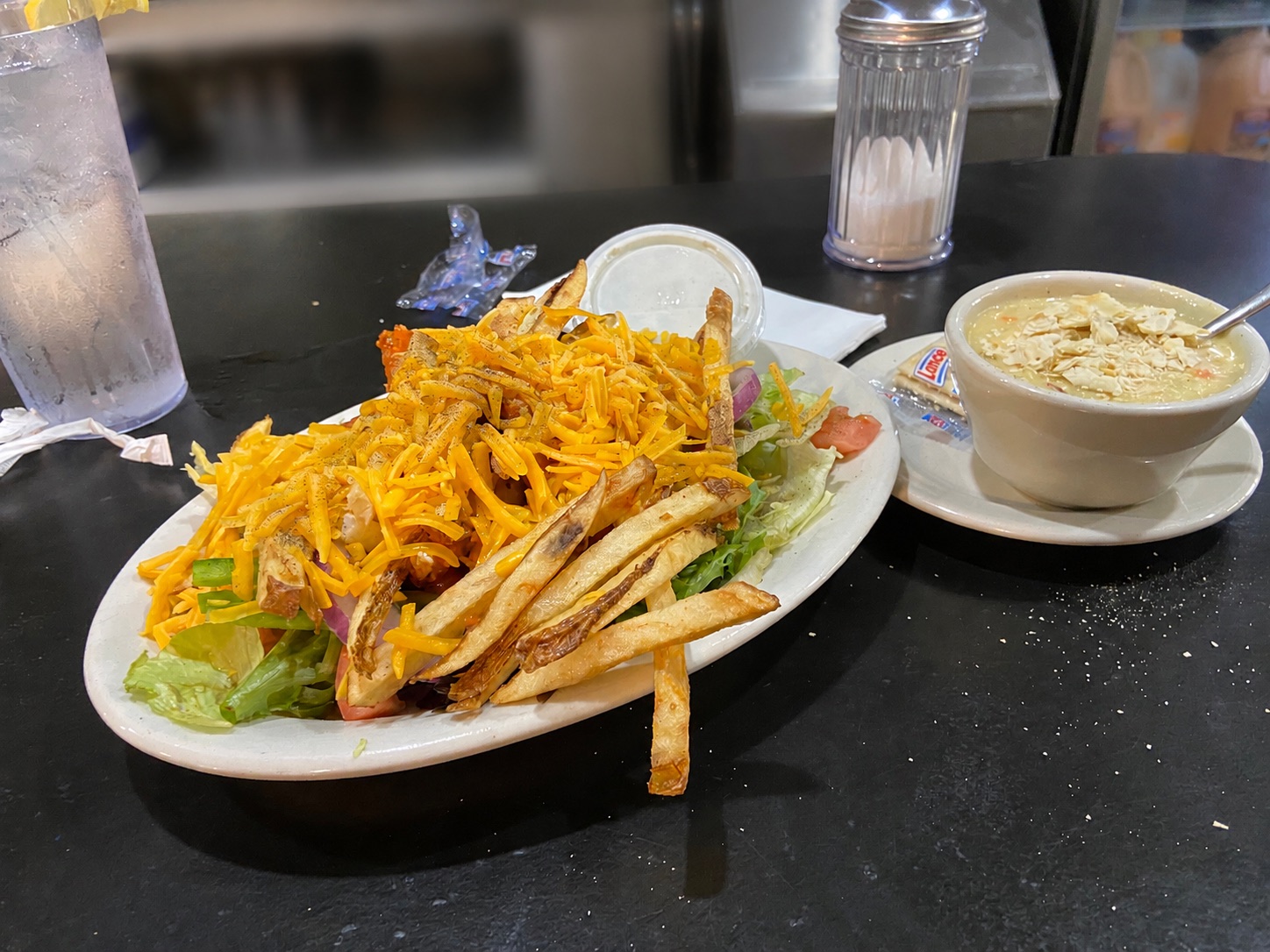 A plate of food featuring a salad topped with shredded cheese and fries, accompanied by a bowl of soup with crackers, sits on a black table alongside a glass of water and a container of sugar packets.